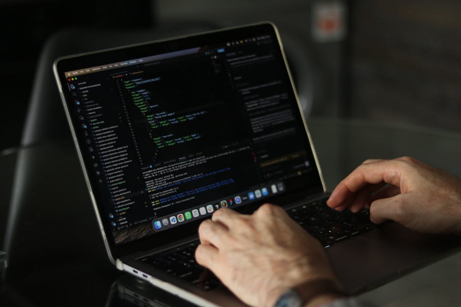 A close-up view of a person’s hands using a laptop with a black keyboard and a sleek metallic body, displaying a code editor with lines of programming code in various colours on the screen. The lapt