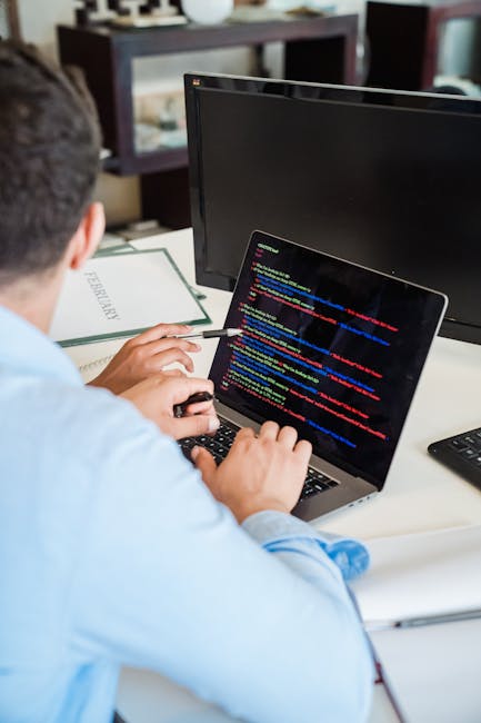 A close-up view of a person’s hands using a laptop with a black keyboard and a sleek metallic body, displaying a code editor with lines of programming code in various colours on the screen. The lapt