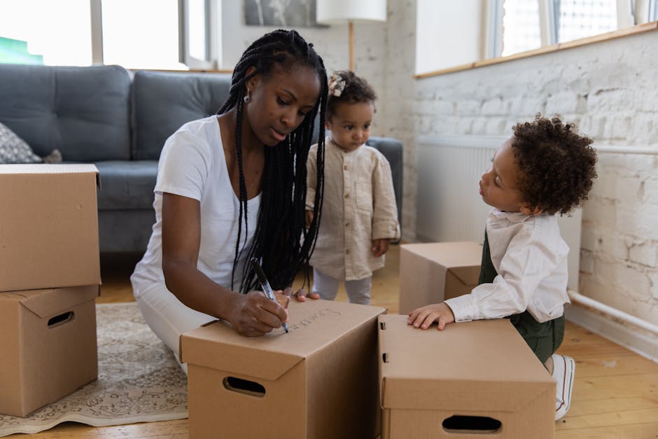 A young girl with curly brown hair, wearing a light pink long-sleeve shirt and grey patterned pants, is standing inside a room surrounded by moving boxes during a home relocation. She is holding a red