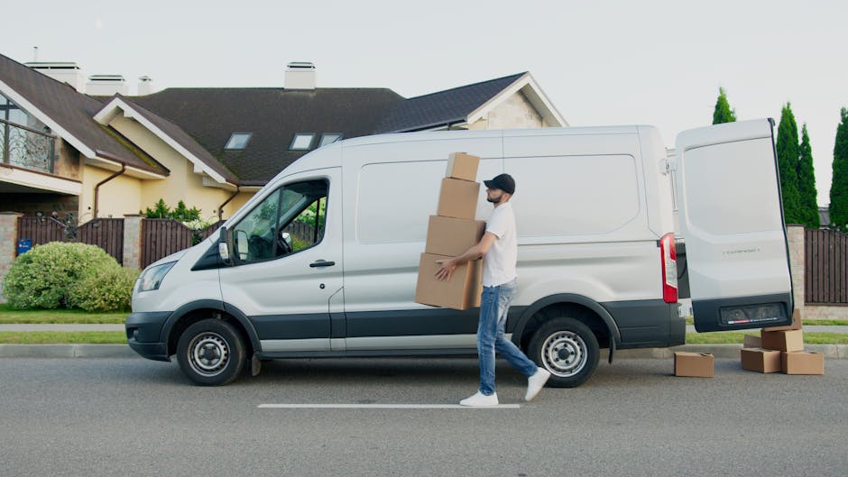 A man dressed in casual clothing, including a white t-shirt, blue jeans, and a black cap, is outdoors next to a white commercial van parked on a residential street. He is carrying three medium-sized c