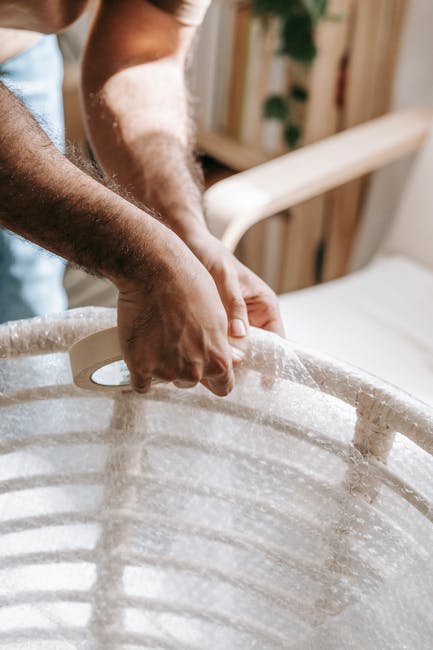 Close-up of a person’s hand carefully wrapping a piece of furniture with protective bubble wrap during a home relocation process. The furniture appears to be a rattan chair with curved wooden slats,