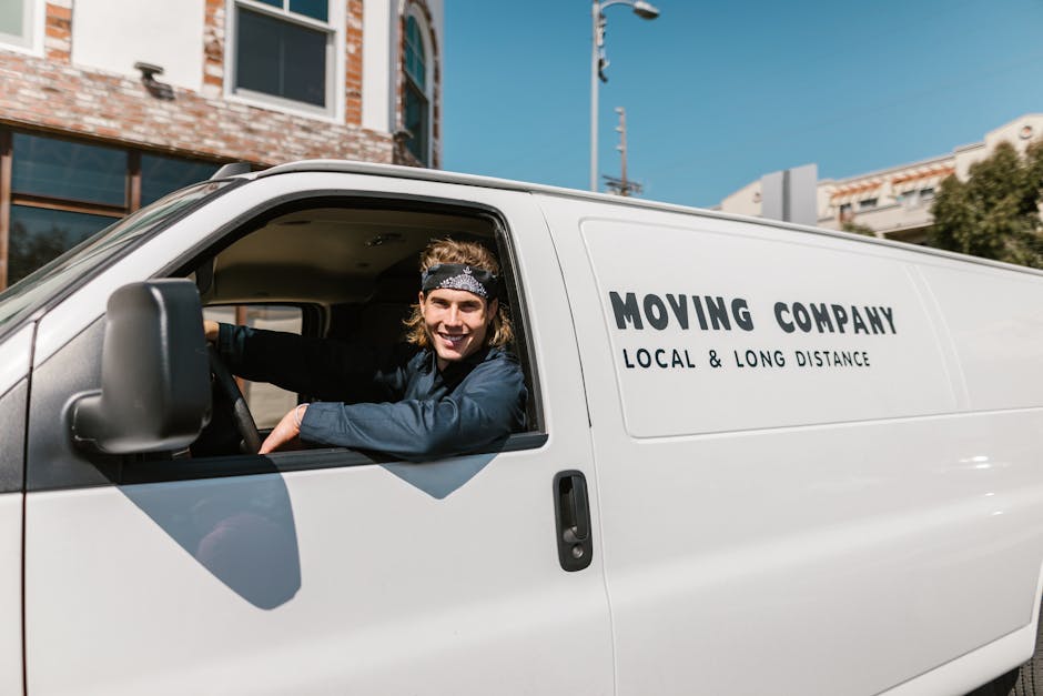 A side view of a white moving van parked on a residential street with a brick building and a no-parking sign visible in the background. The van features the company name 'MOVING COMPANY' and the descr