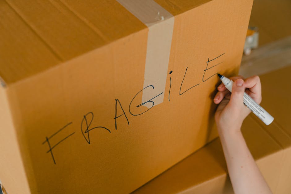A person is writing the word 'FRAGILE' on a large cardboard box with a black marker, indicating careful handling during a home relocation or furniture transport process. The box is positioned inside a